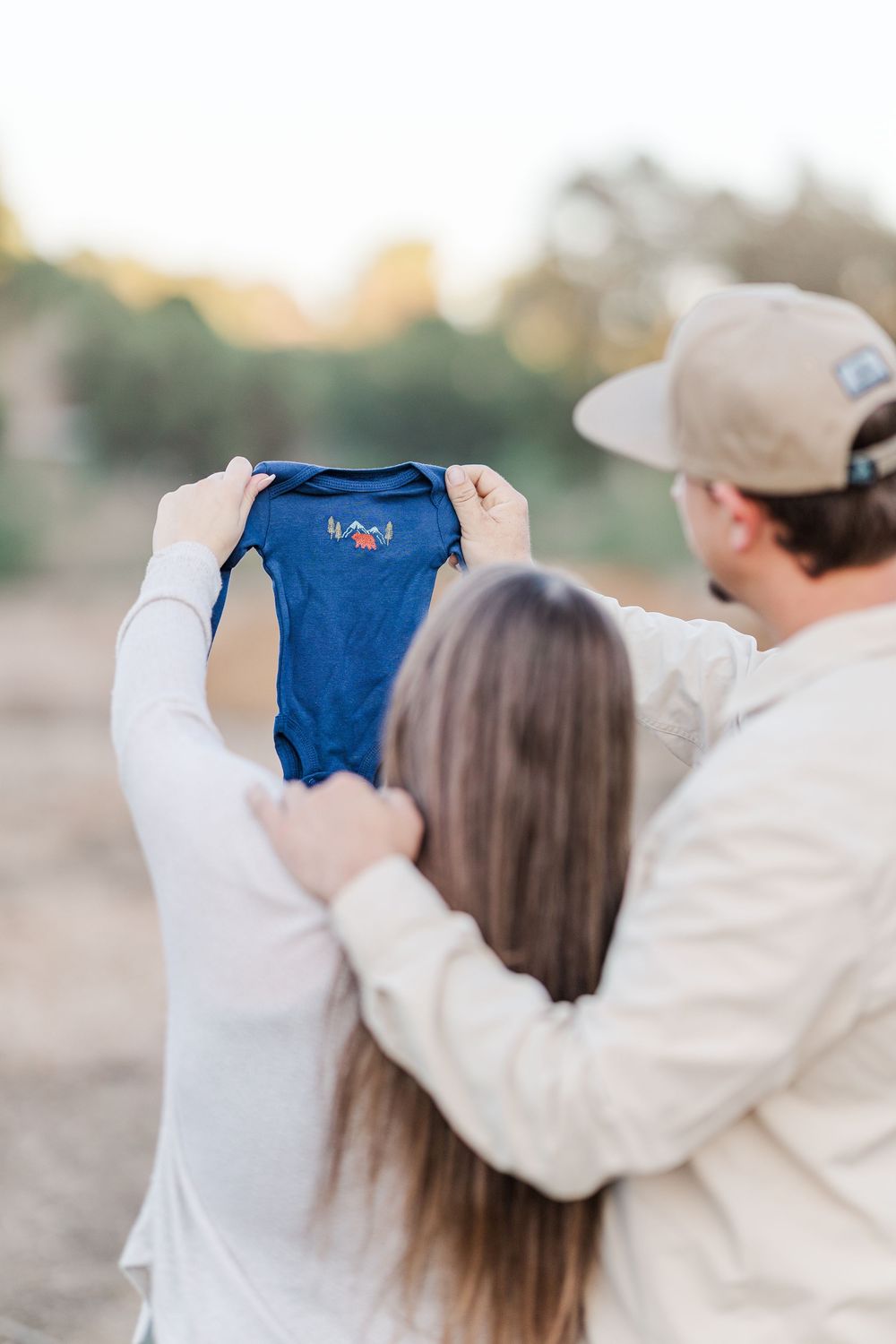 A couple in white outfits holding up a small blue baby onesie together against a soft sunset background.