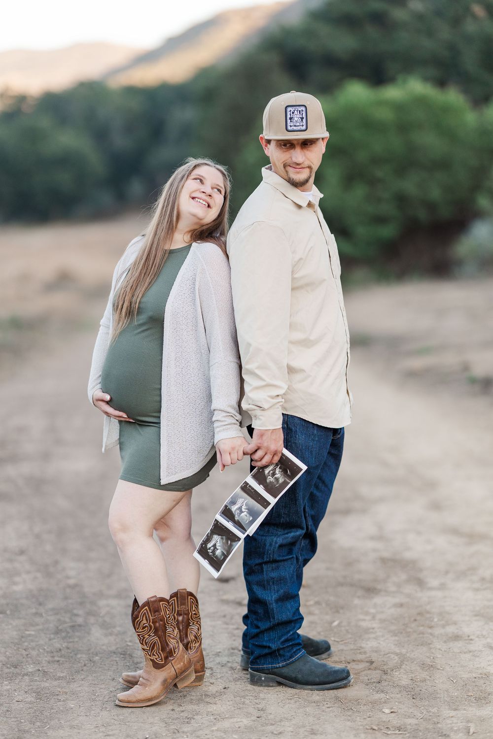 Expecting couple poses outdoors holding ultrasound photos, with her in cowboy boots and a green dress.