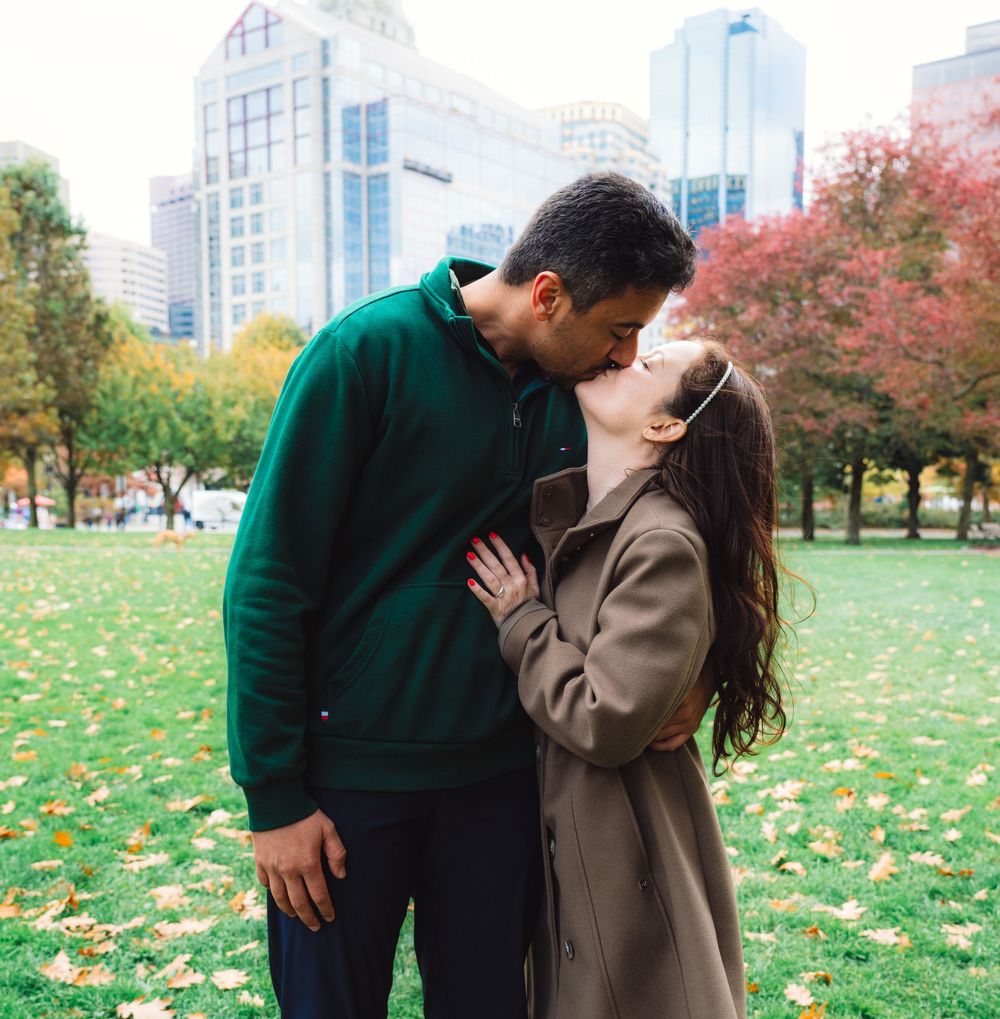 Newly engaged couple embracing in a city park with fall foliage and skyscrapers in background.