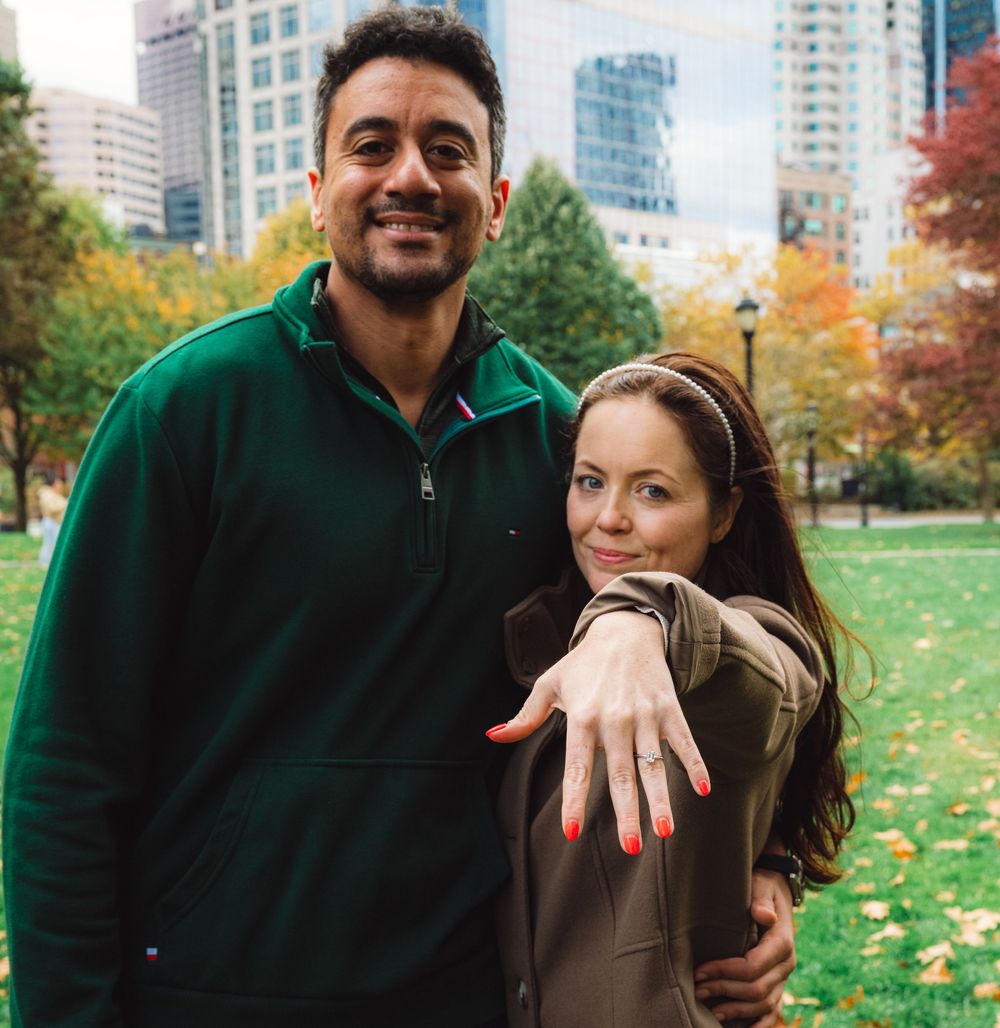 Happy couple showing off engagement ring in urban park during fall season.
