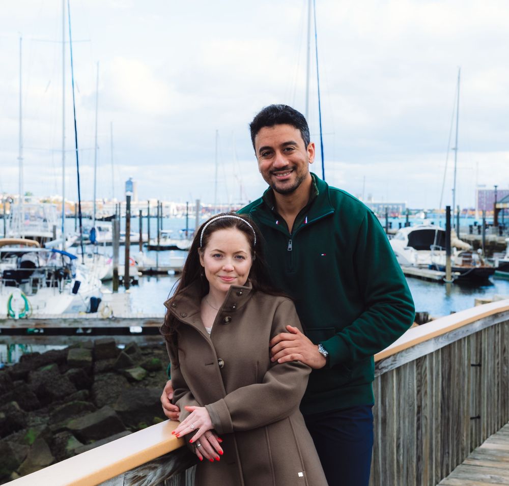 Engaged couple posing at marina dock with boats and cloudy sky in background.