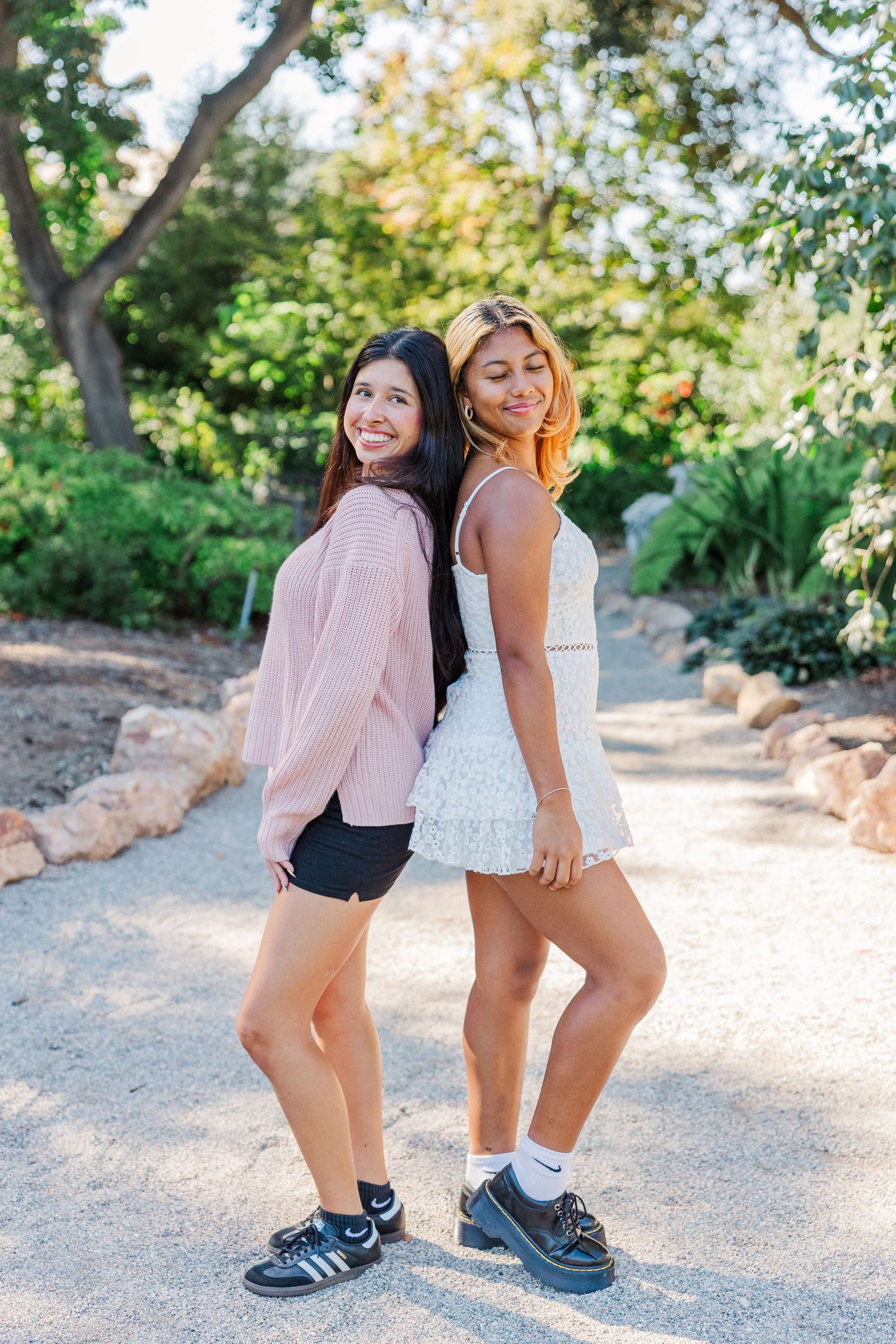 Two people wearing casual outfits and sneakers stand together on a sunny dirt path surrounded by greenery.
