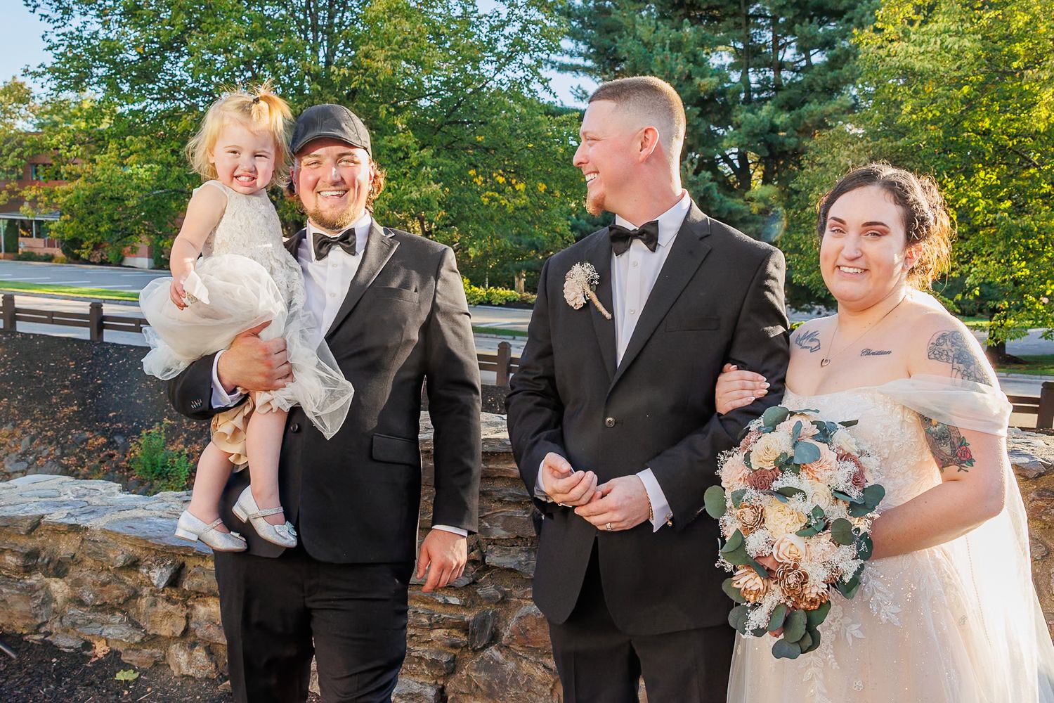 Wedding party in formal attire gathers outdoors near stone wall with lush greenery in background.