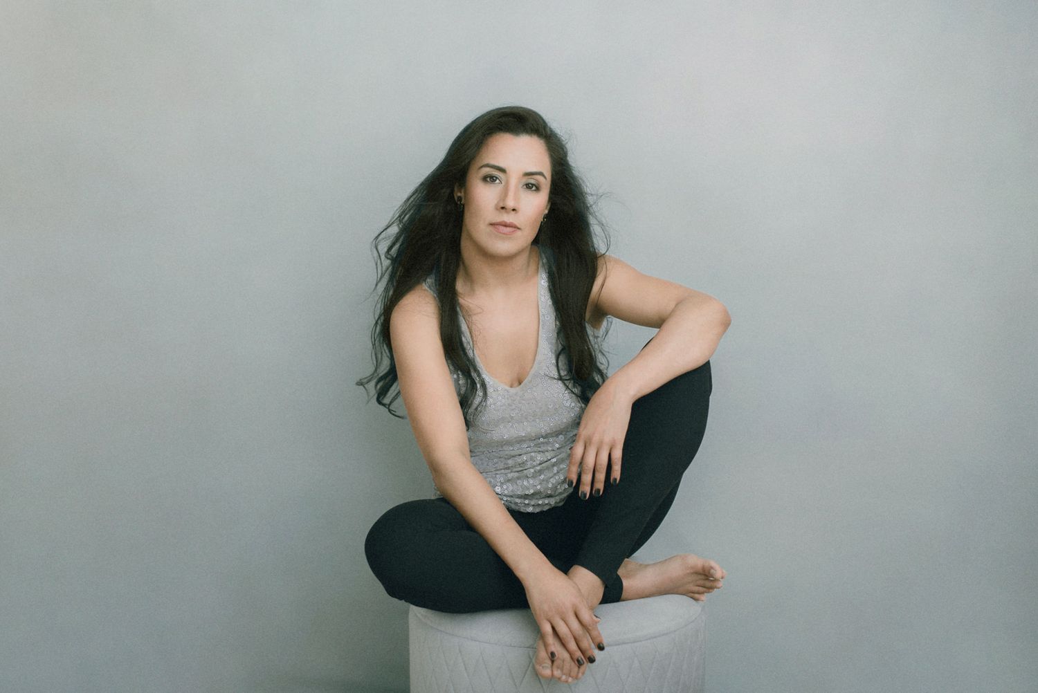 A person in a gray tank top and black pants sits casually against a gray backdrop in a studio setting.