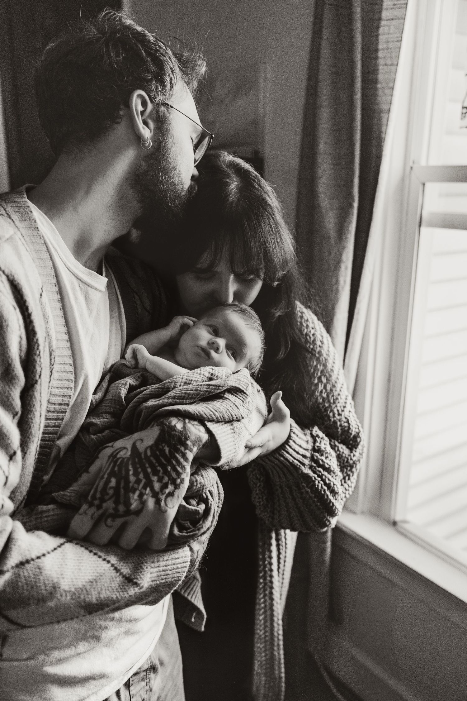 Black and white portrait of new parents lovingly kissing their sleeping newborn by a window.
