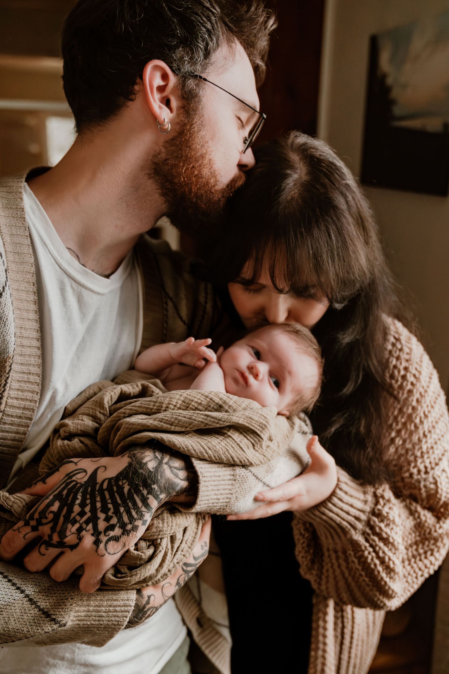 Loving parents kissing their peaceful newborn baby held in fathers tattooed arms indoors.