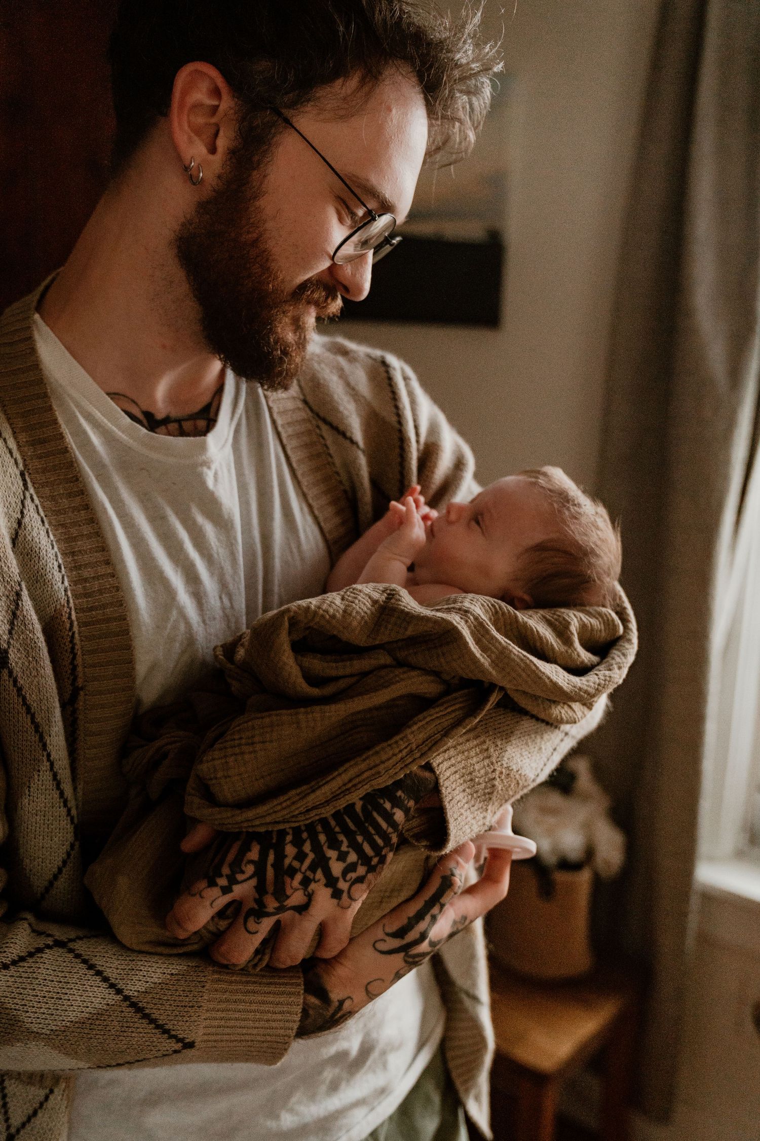 Bearded tattooed father in glasses gazing lovingly at his newborn baby in a warmly lit room.