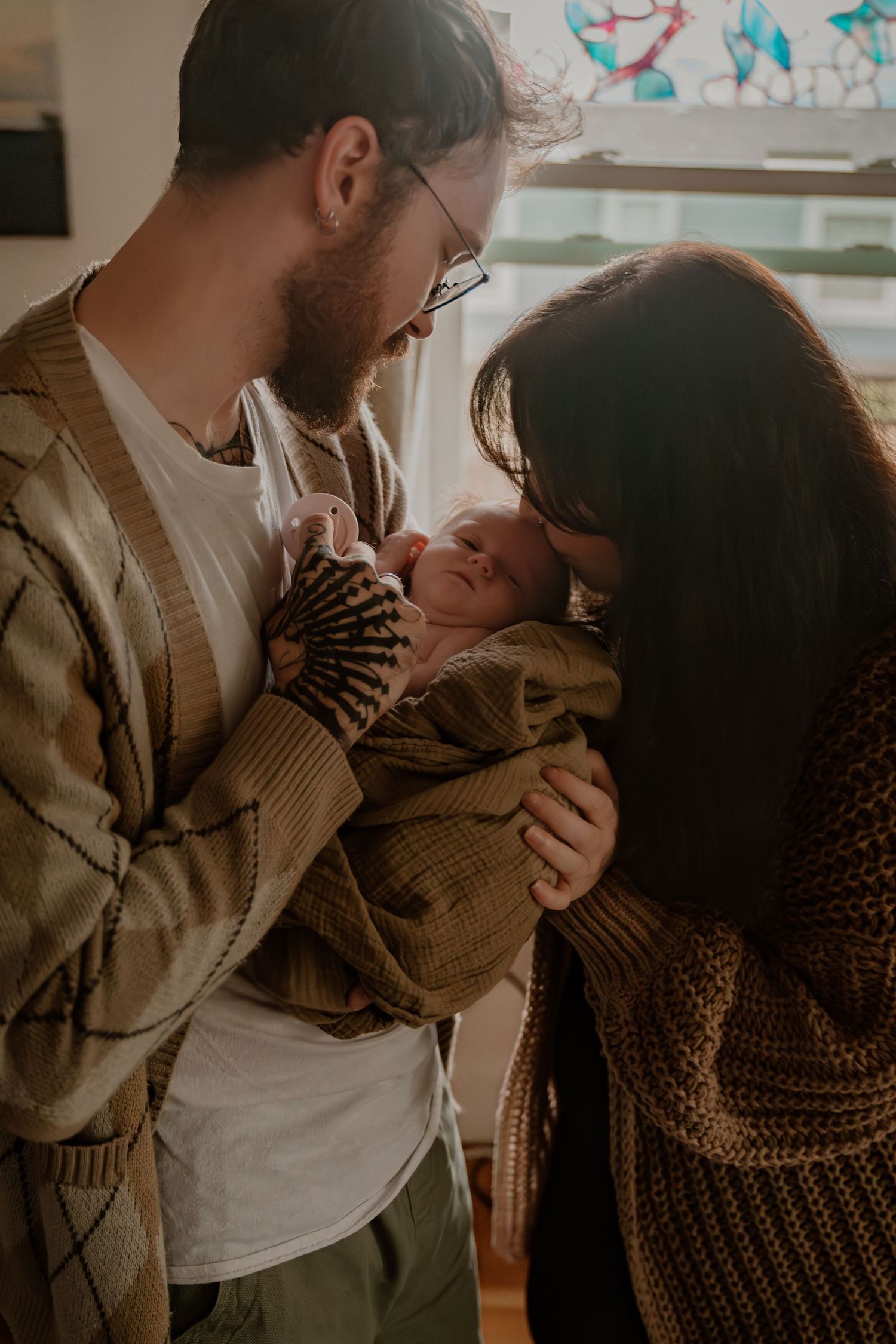New parents lovingly gaze at their sleeping newborn baby wrapped in a cozy knit blanket by a sunlit window.
