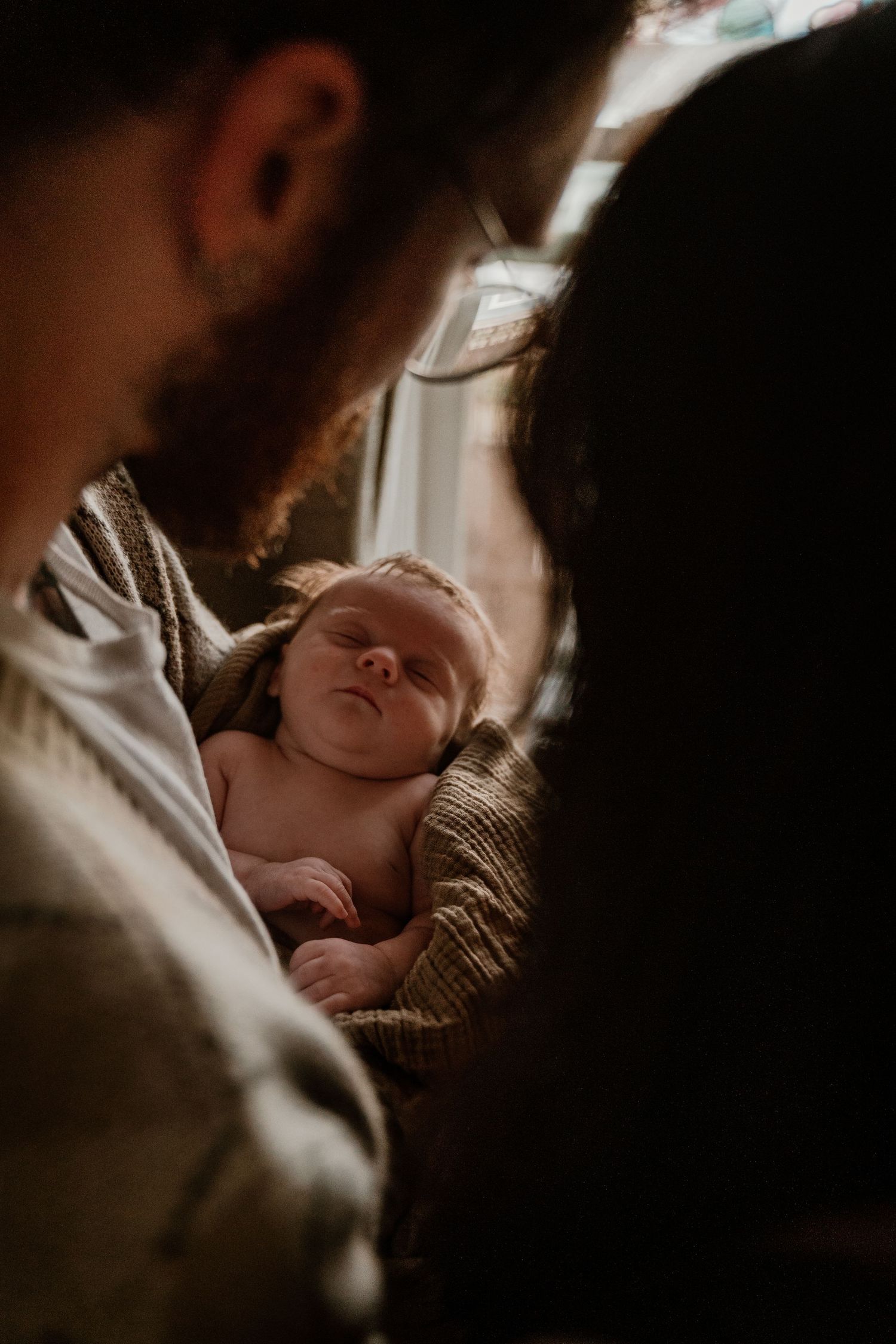 Overhead view of a peaceful newborn baby sleeping between two parents in warm natural light.