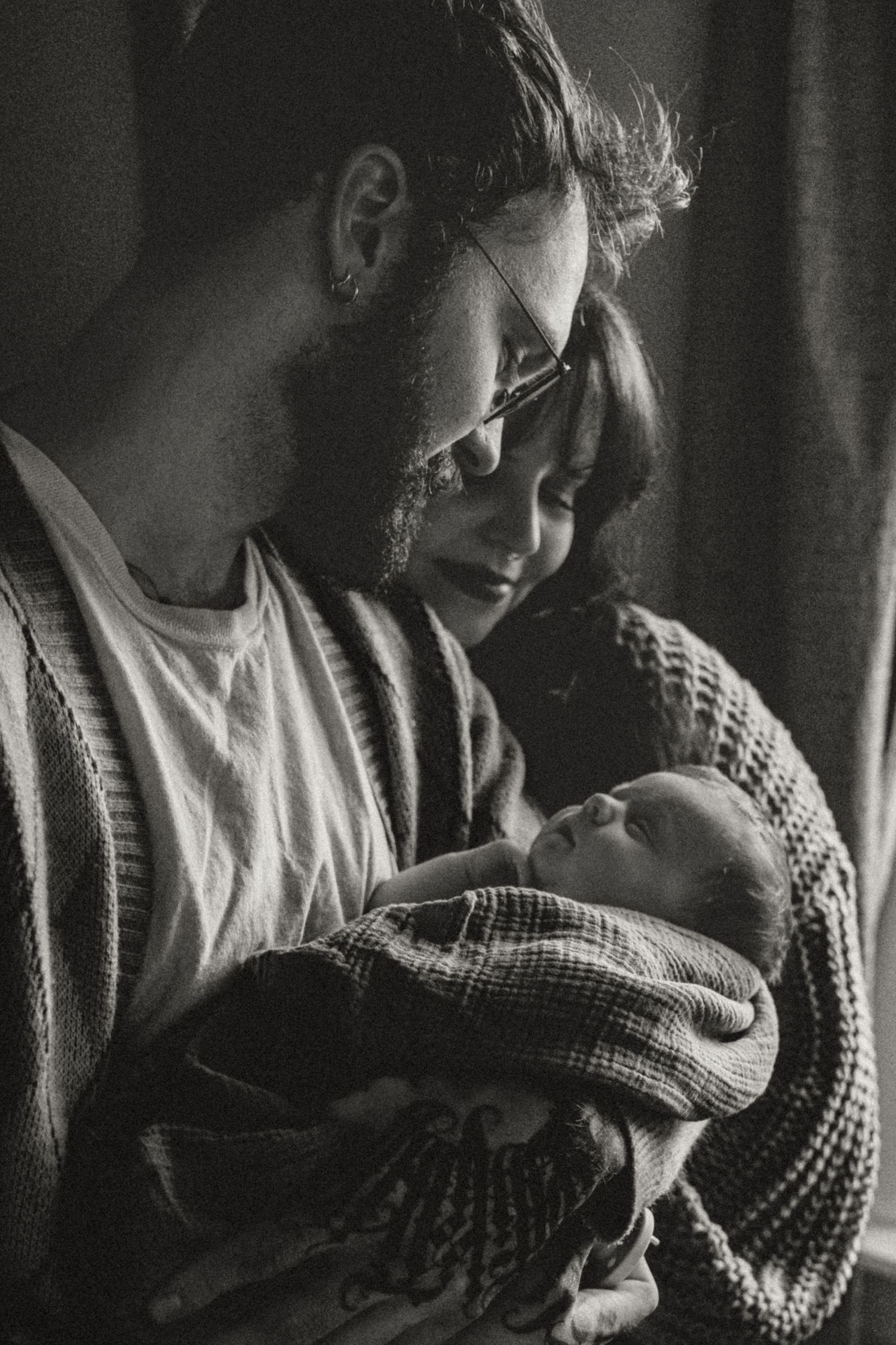 Black and white photo of new parents tenderly holding their swaddled newborn baby together.