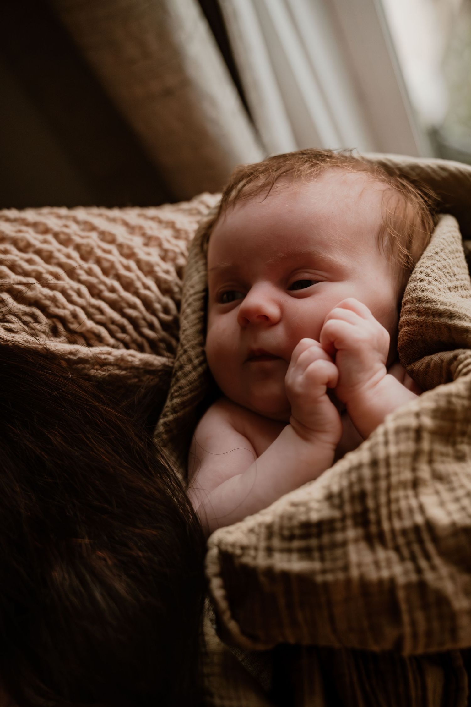 Awake newborn baby with dark hair lying on a brown knit blanket, gazing upward with tiny fist near face.