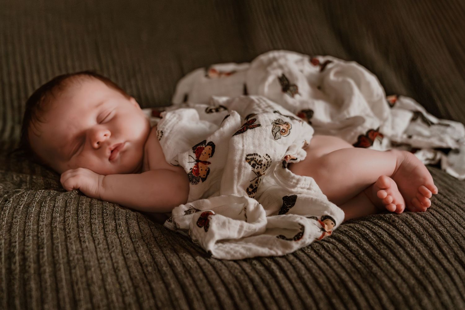 Sleeping newborn baby wrapped in a white butterfly print muslin swaddle on a green knit surface.