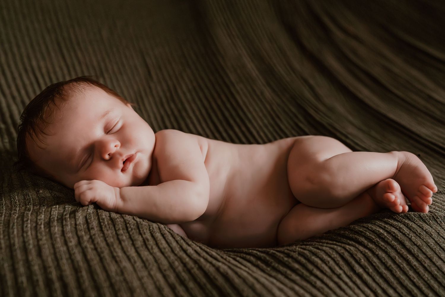 Peaceful naked newborn baby sleeping curled up on a soft olive green knit blanket.