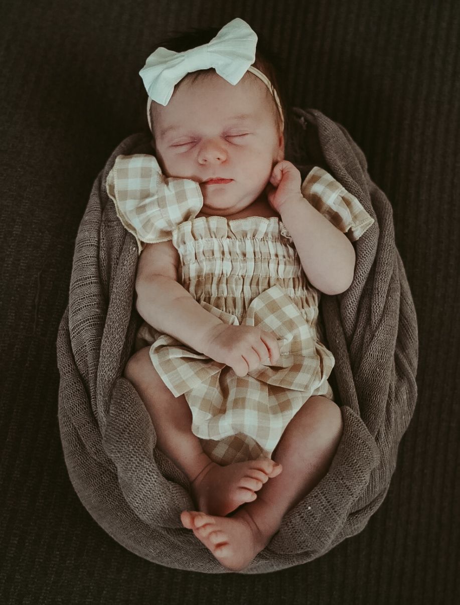 Sleeping newborn baby girl in a gingham smocked romper and white bow headband in a round basket.