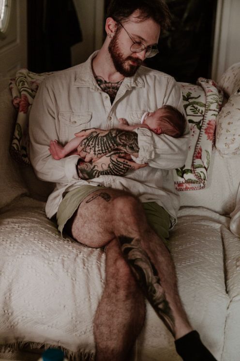 Tattooed father sitting in a vintage armchair cradling his sleeping newborn baby against his chest.