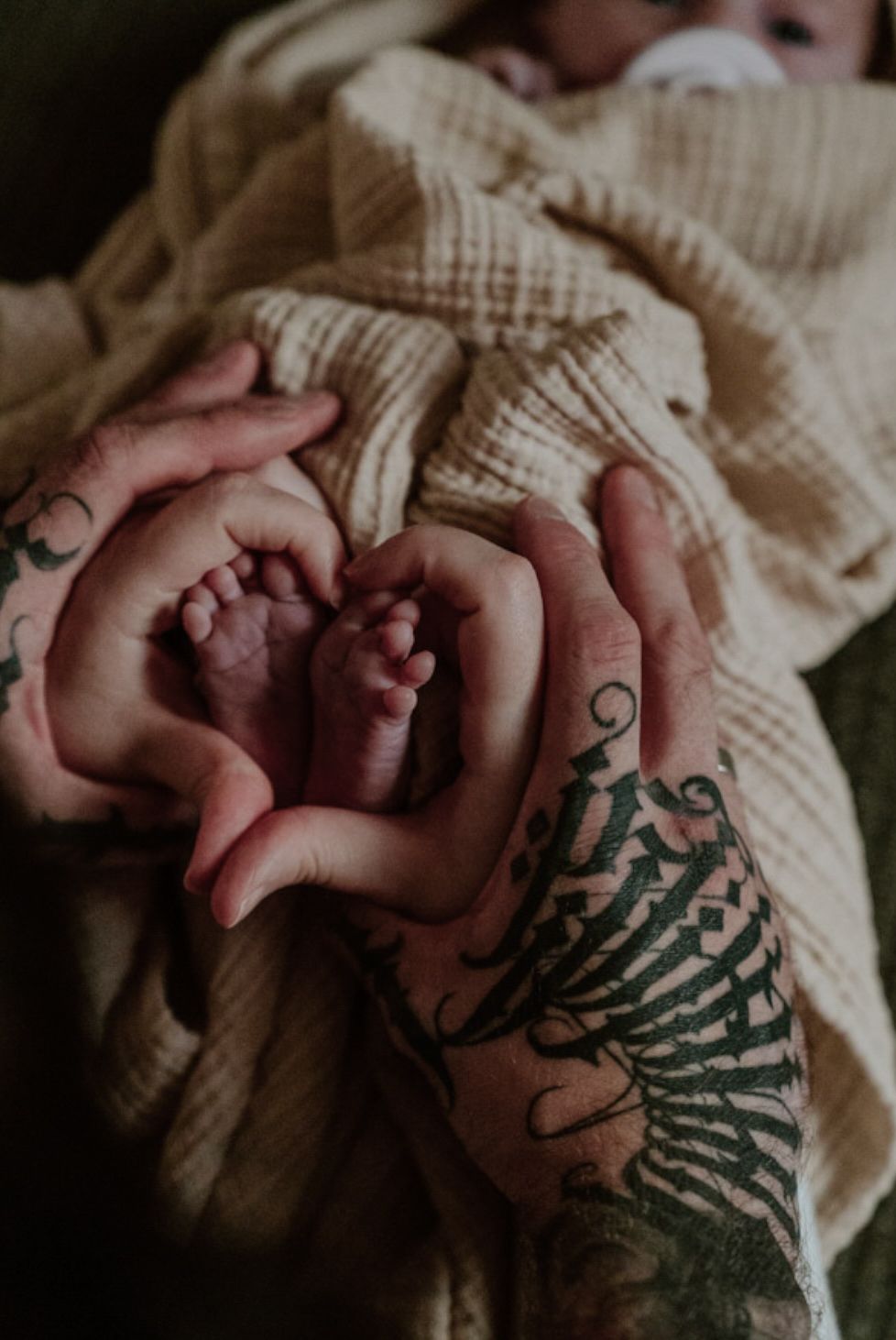 Two tattooed parents cradle their newborn baby's tiny feet in a heart shape with a cream blanket.