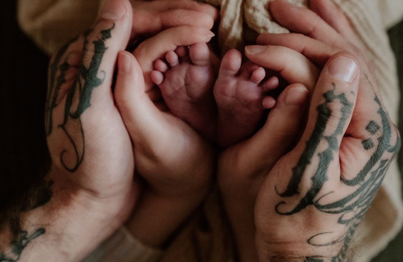 Close-up of two tattooed hands gently cupping a newborn baby's tiny feet together.
