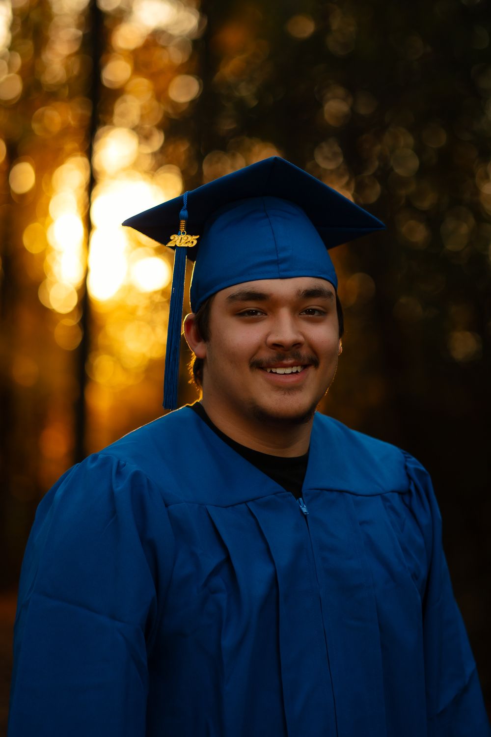 Graduate in royal blue cap and gown smiles warmly against a golden sunset bokeh background.