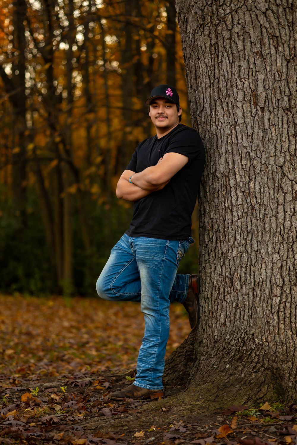 A person in a black shirt and jeans leans casually against a large tree trunk in an autumn forest setting.