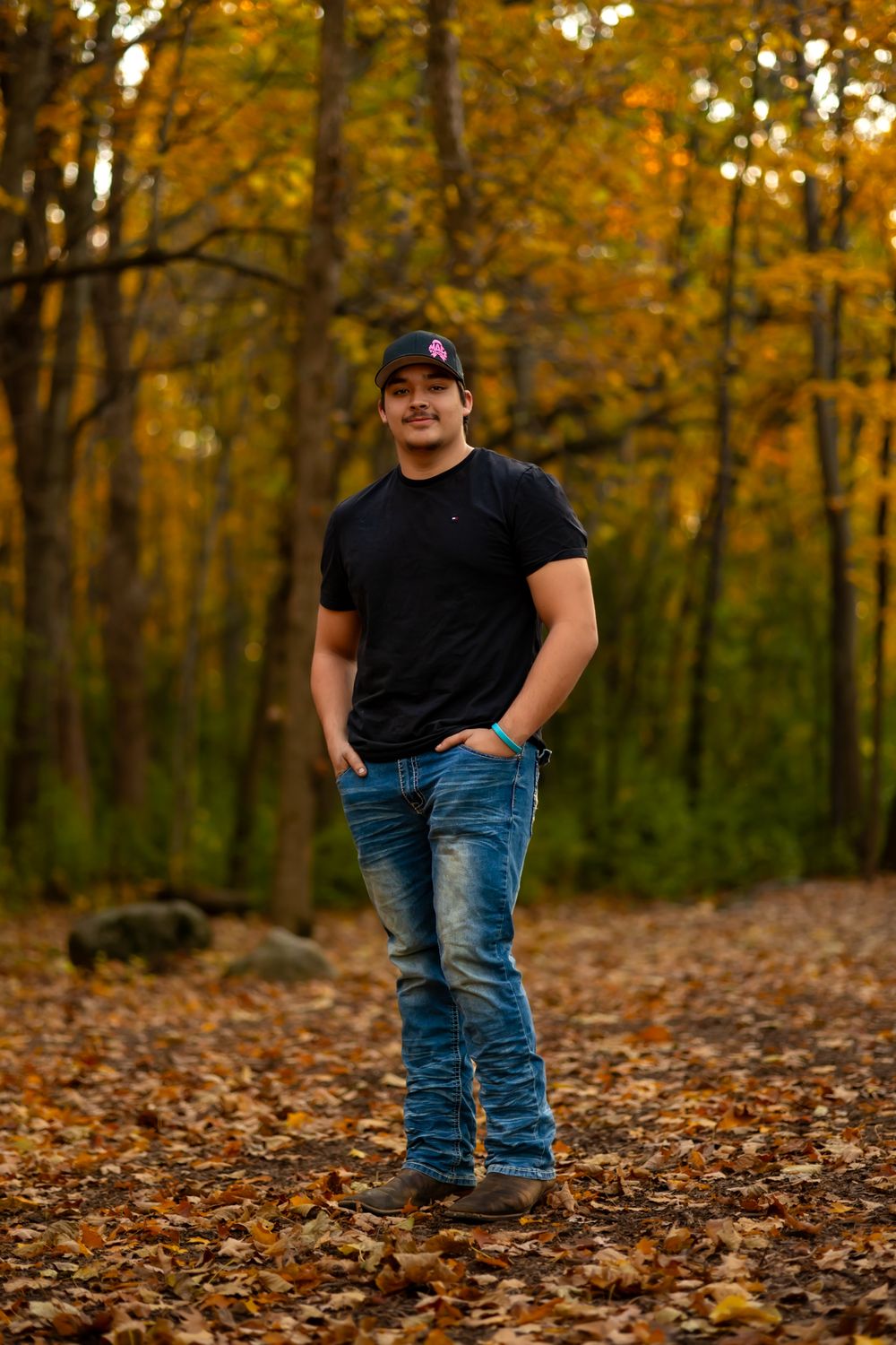 A person wearing a black t-shirt and blue jeans stands on fallen autumn leaves in a forest with golden foliage.