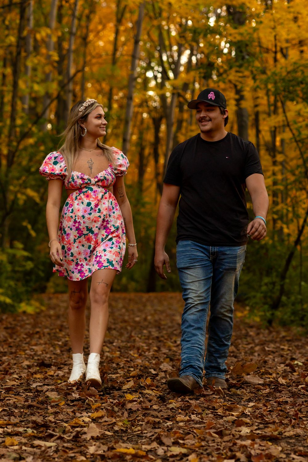A couple walks through fallen autumn leaves in a forest with golden foliage, wearing casual attire.