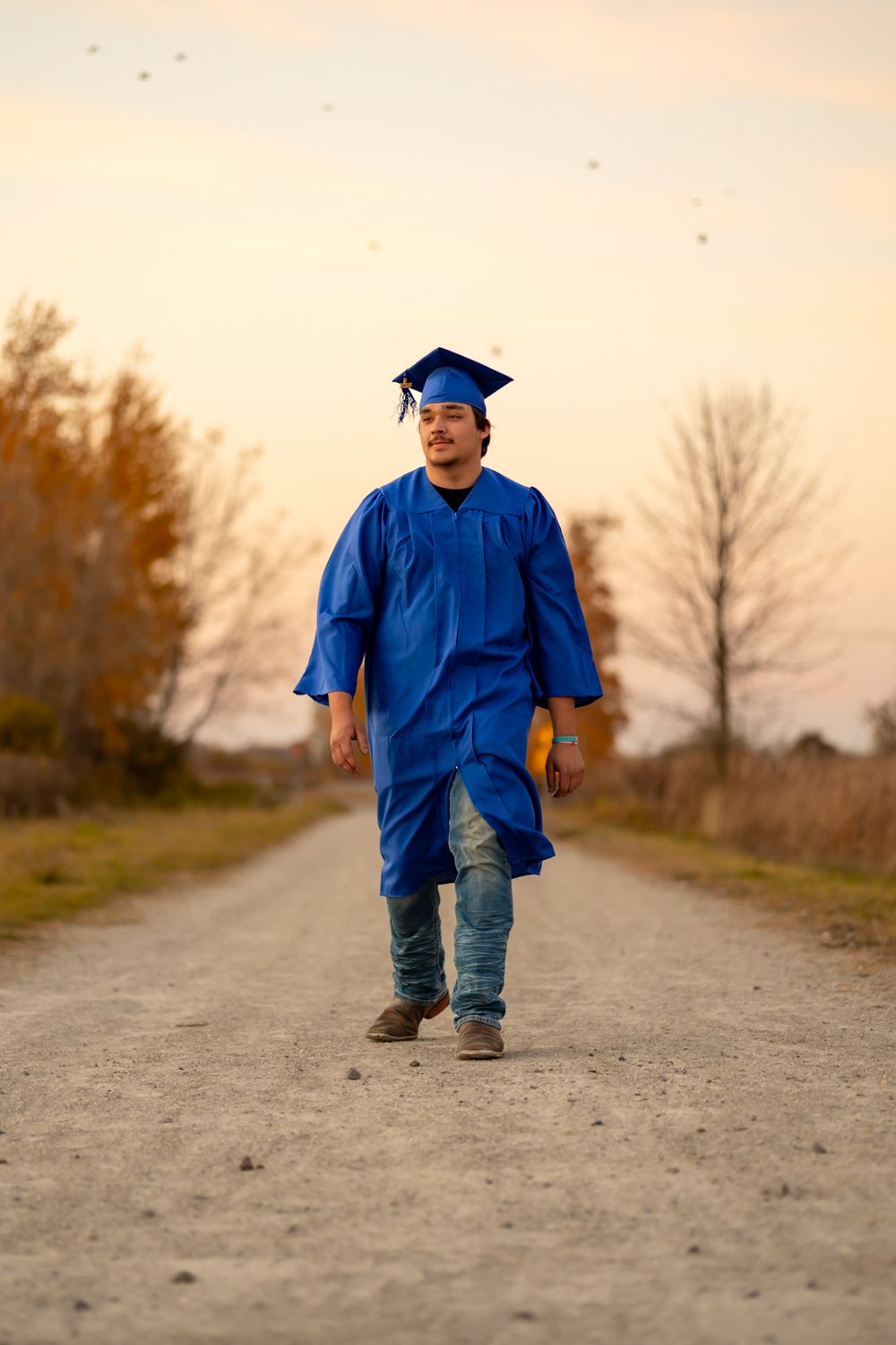 Small figure in blue graduation cap and gown walks away on dirt path during golden hour.