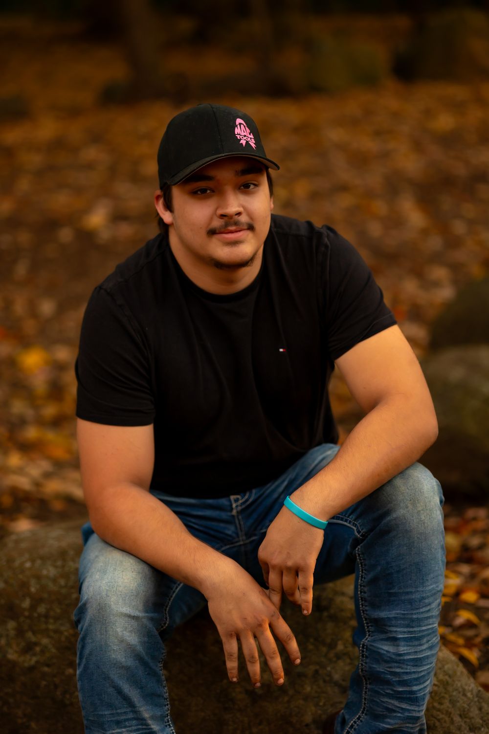 A person in a black t-shirt and baseball cap sits outdoors against a backdrop of autumn trees.