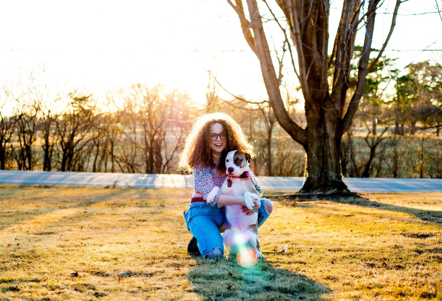 A person with curly hair sits outdoors in jeans during golden hour as long tree shadows stretch across the ground.