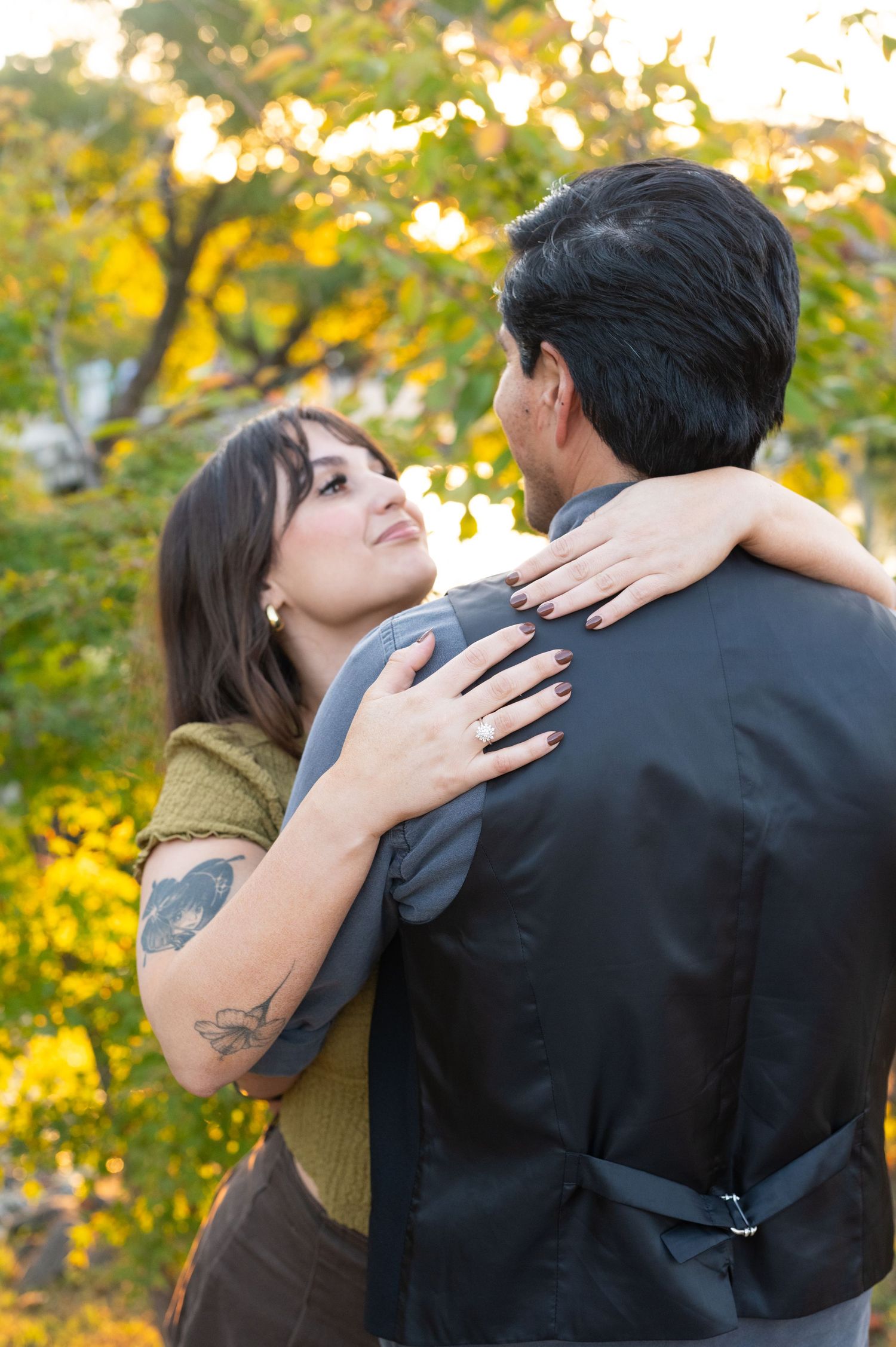 Woman with tattoos embracing man while showing engagement ring in autumn park.