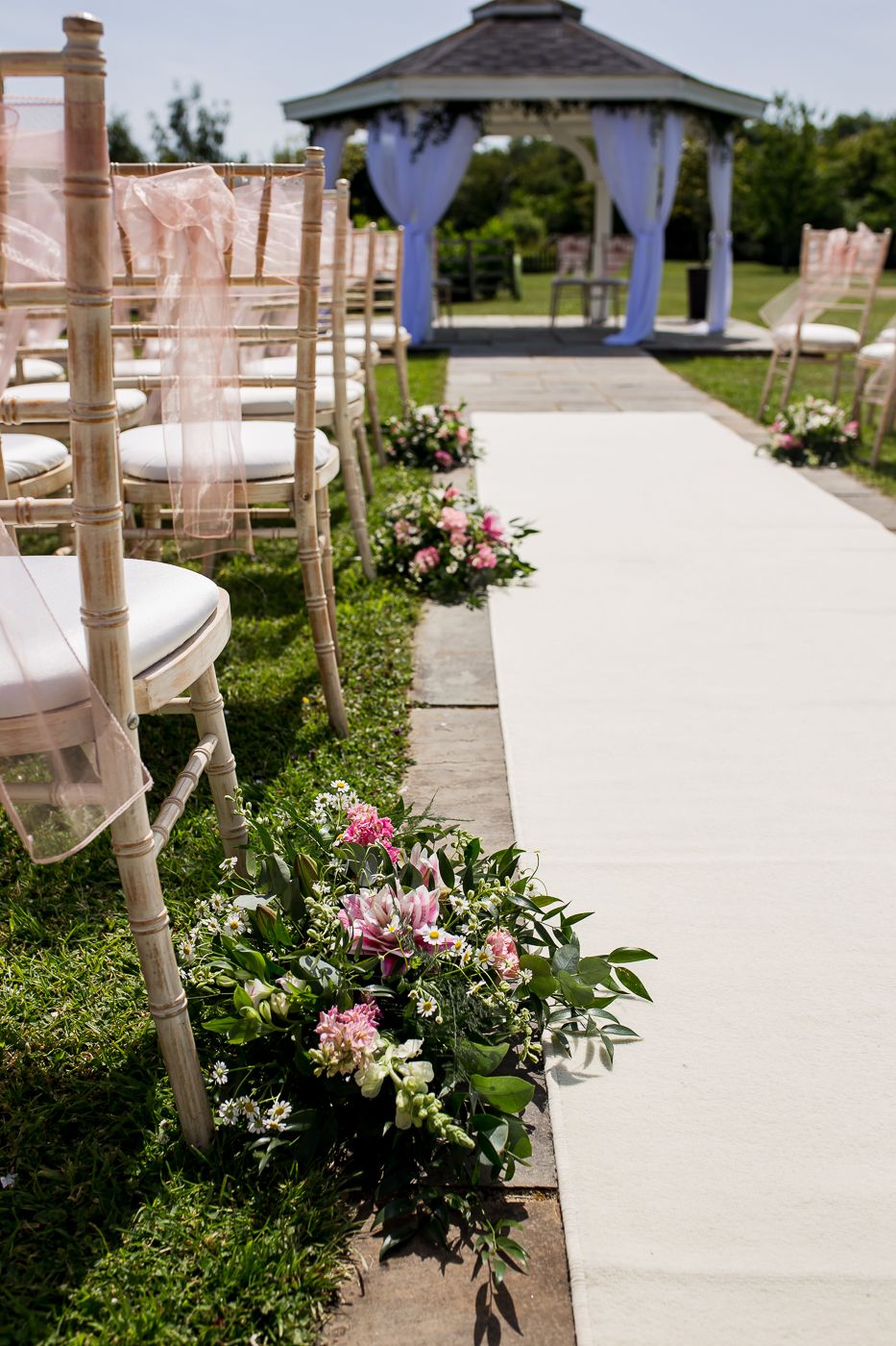 White aisle runner lined with pink floral arrangements leads to outdoor wedding gazebo.