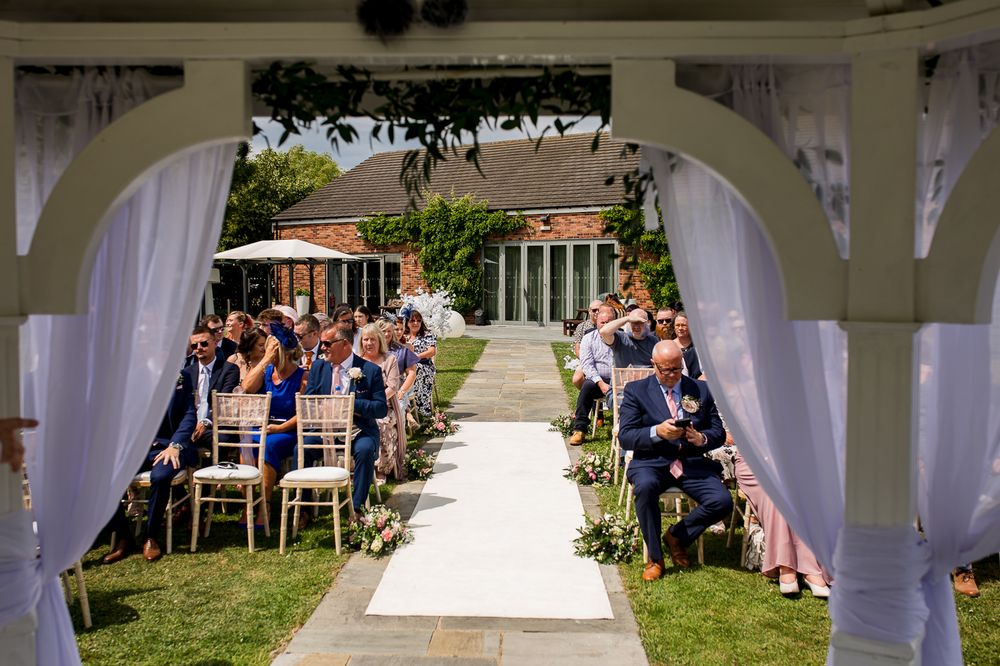 Wedding guests seated in white chairs on a lawn during an outdoor ceremony under white fabric draping.