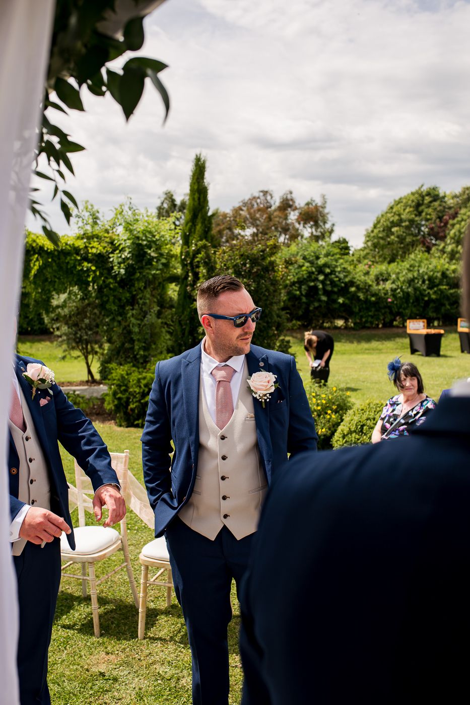 Groomsmen in navy suits and beige vests stand under white pergola during outdoor wedding.
