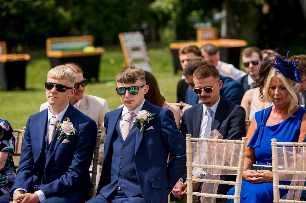 Wedding guests in formal attire seated on wooden chairs during an outdoor ceremony.