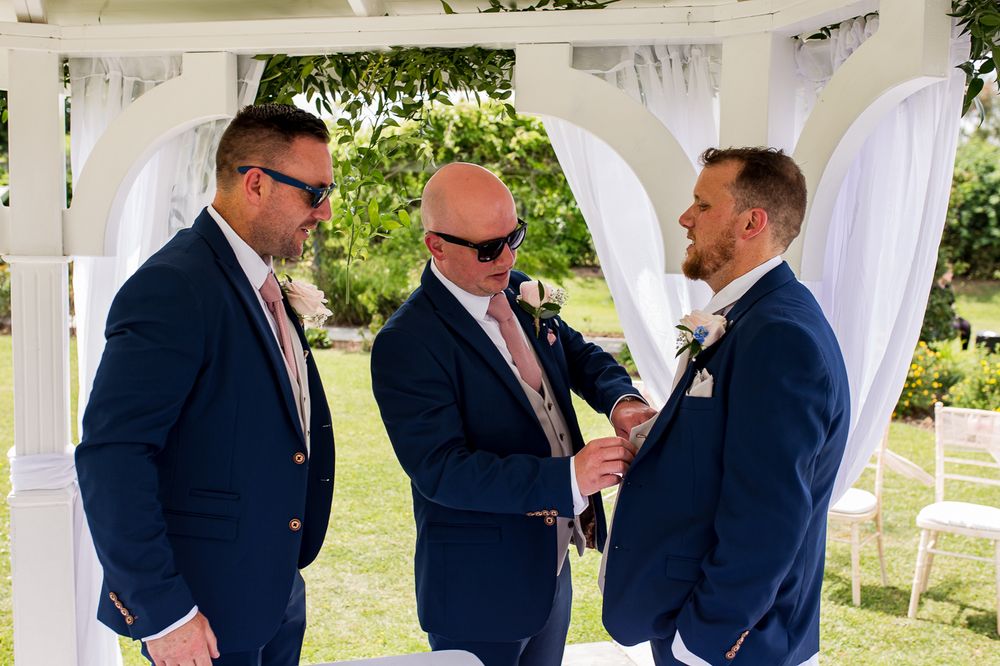 Wedding party members in navy suits gather under a white pergola during an outdoor celebration.