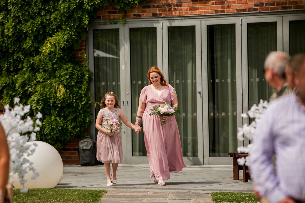 Bridesmaids in pink dresses walk along brick building exterior during wedding celebration.