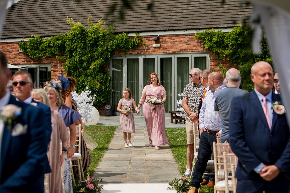 Wedding guests mingle outside a brick building with pink-dressed attendants walking past.
