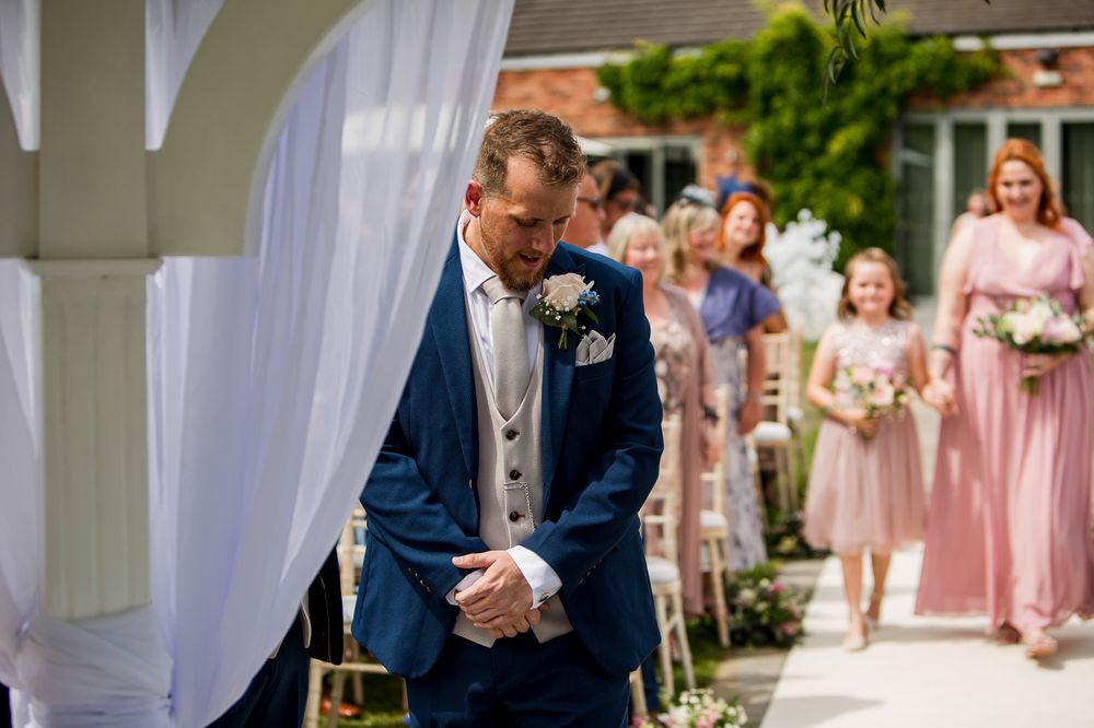 Wedding party in pink bridesmaids dresses and navy suits gather at outdoor ceremony venue with white draping.