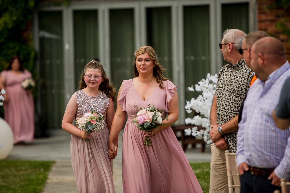 Wedding party members and guests mingle outside brick venue with pink-clad bridesmaids visible.
