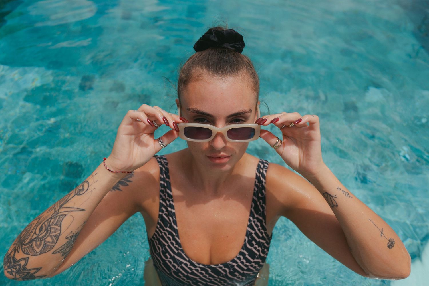 Person in black and white swimsuit enjoying pool water with sunglasses and visible tattoos.