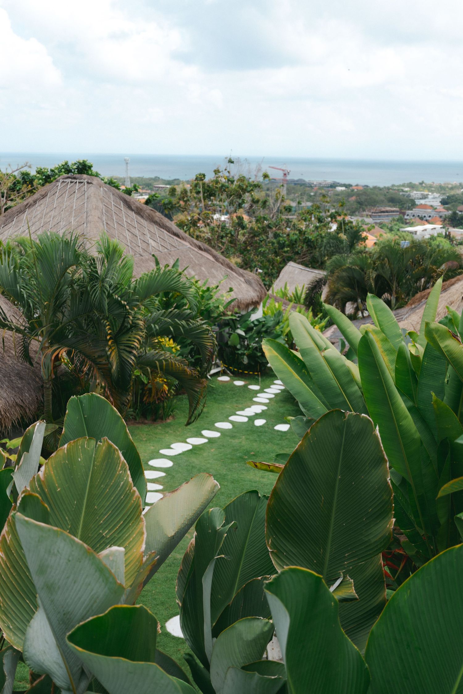 Lush tropical garden with stone pathways and thatched roof structures overlooking an ocean view.