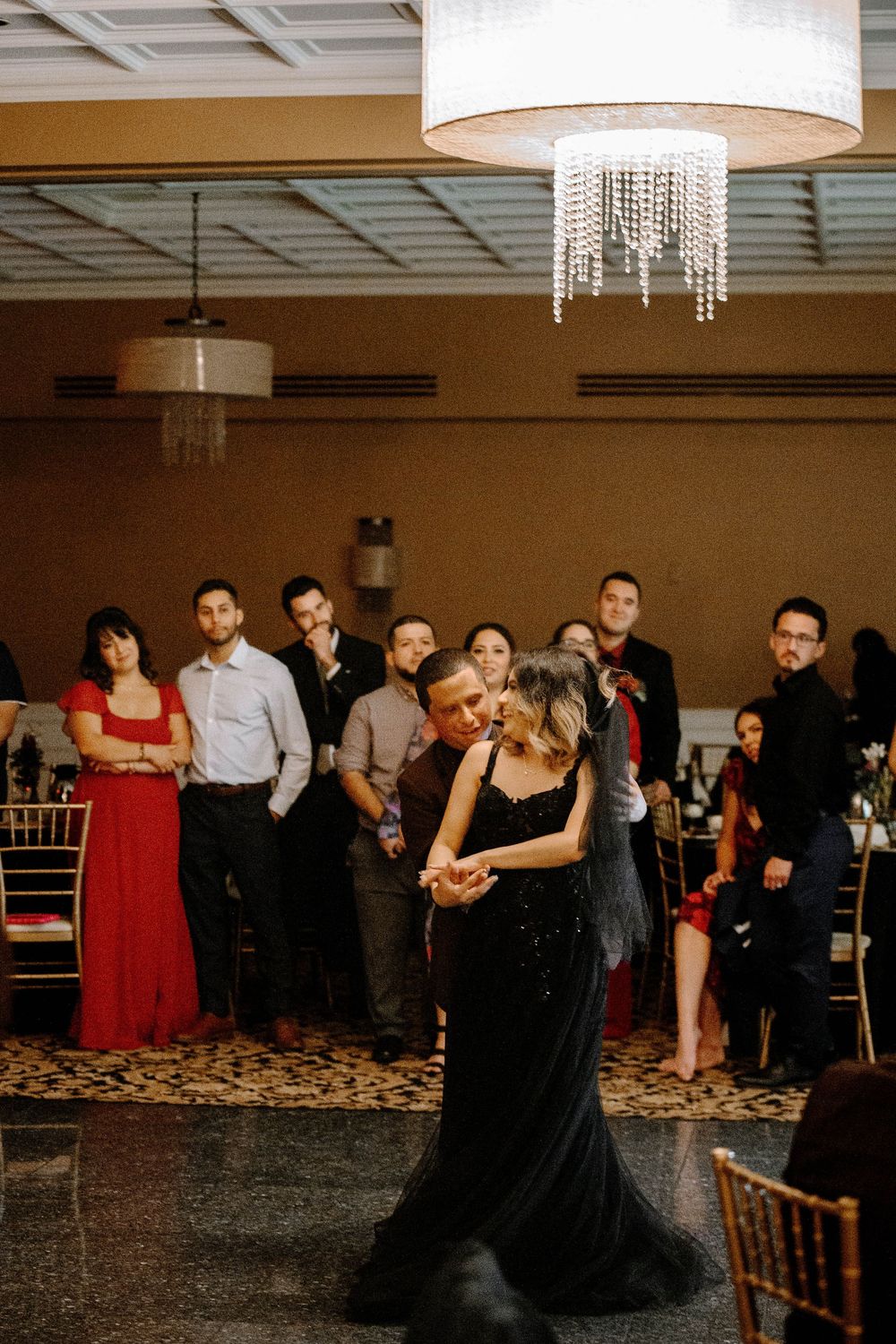 Couples dancing together at an elegant reception venue with crystal chandeliers and wooden chairs.