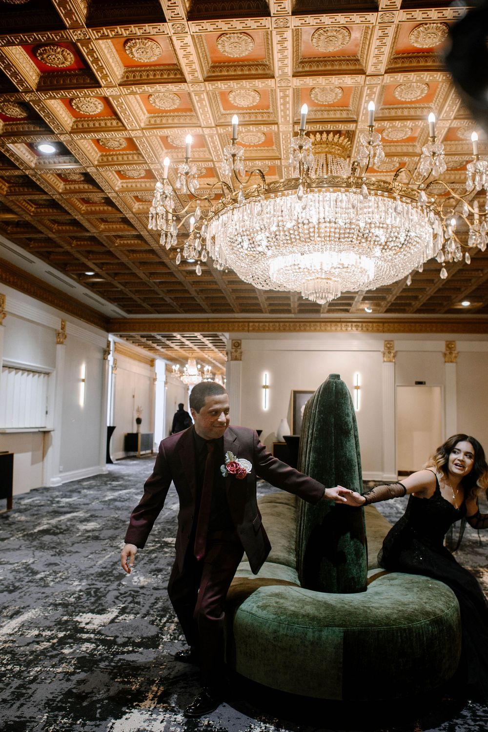 Elegant ballroom with ornate chandeliers and a luxurious green velvet chaise lounge against dark flooring.