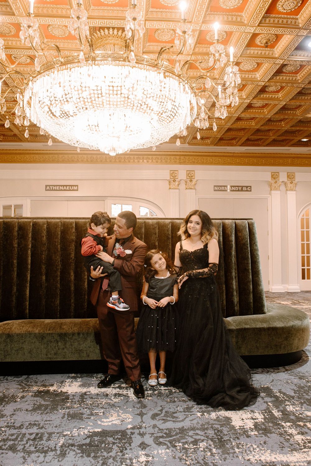 Wedding party poses playfully in an elegant ballroom with crystal chandeliers and ornate decor.