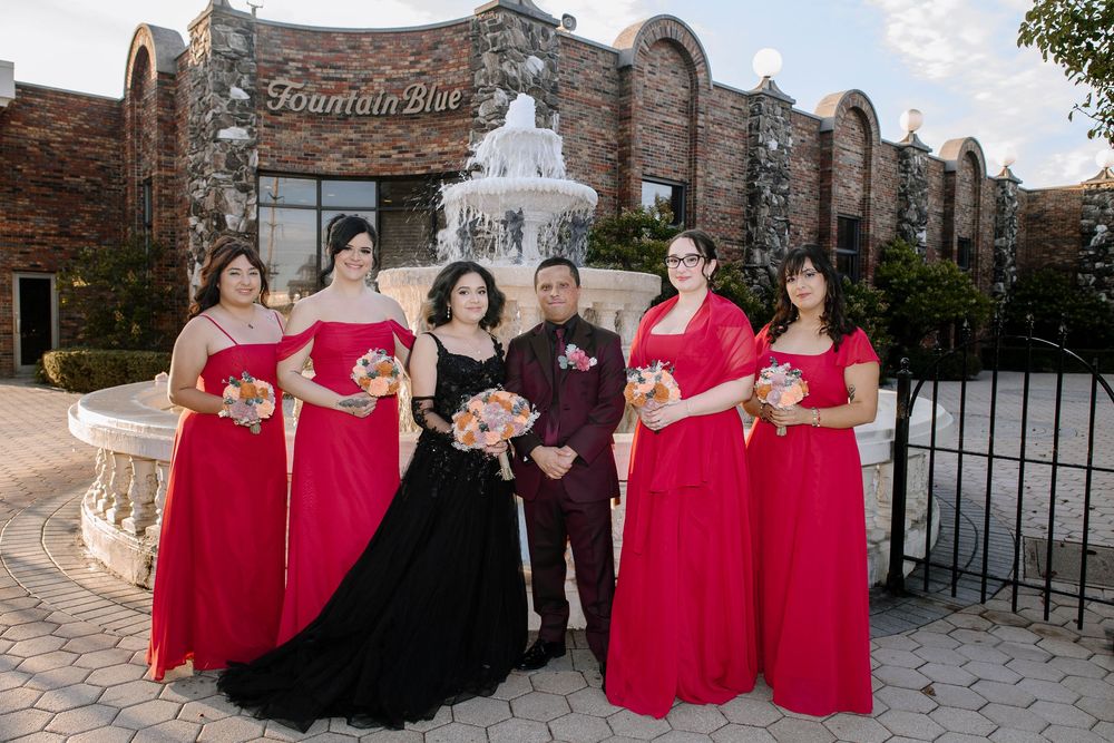 Wedding group in red and black formal wear stands by stone architecture during sunset.