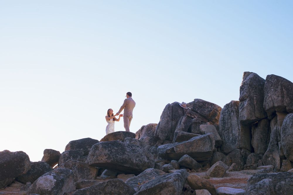Couple holding hands atop rocky boulders against clear sky at sunset wedding photo.