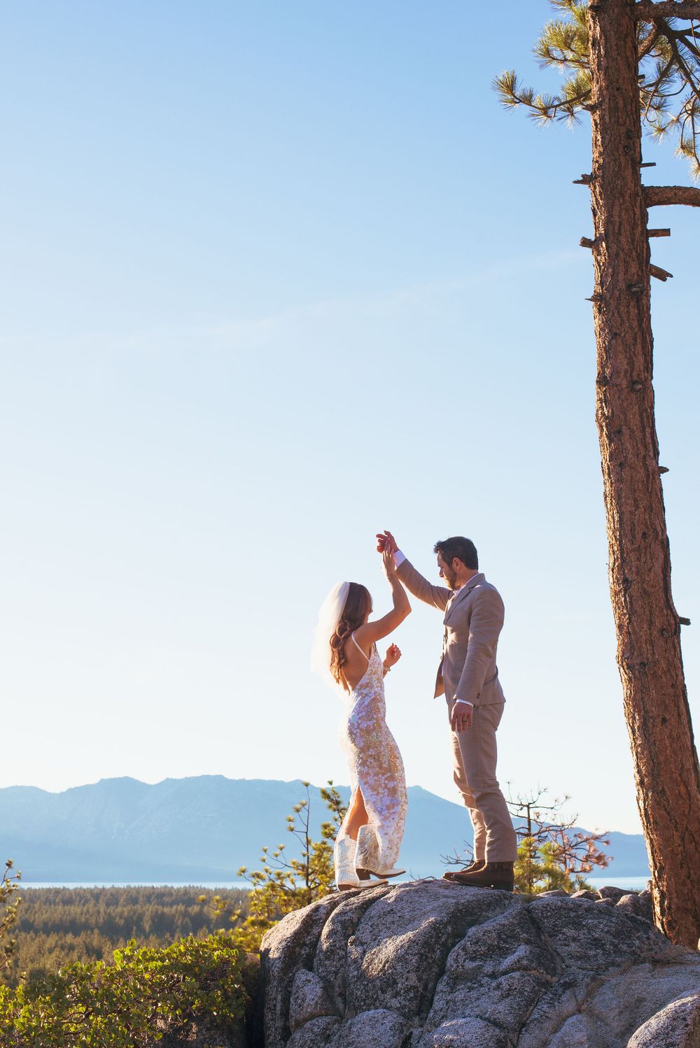 Bride and groom dancing on rocky cliff with pine tree and mountain lake view.