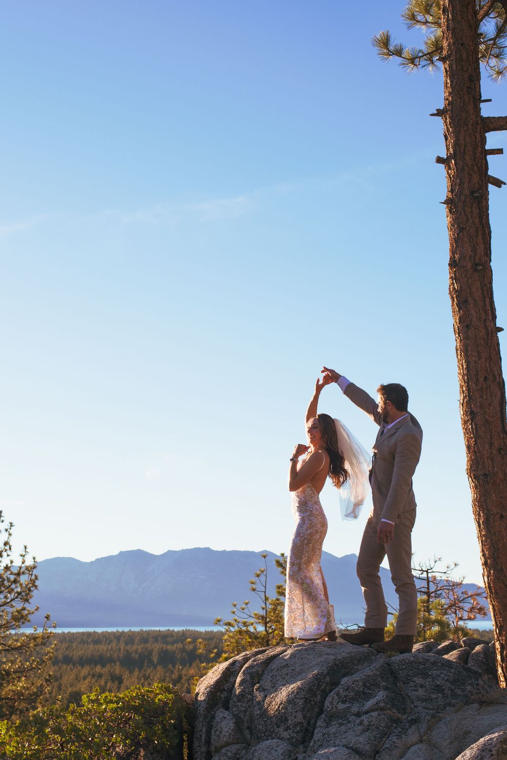 Groom twirling bride in veil on rocky overlook with Lake Tahoe mountain backdrop.