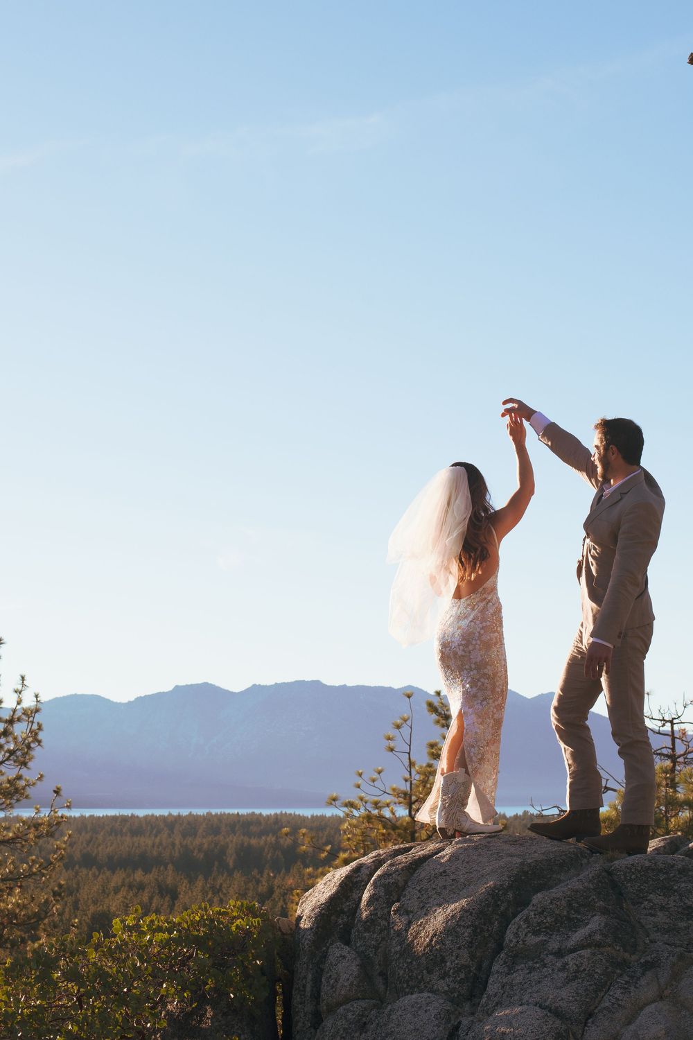 Bride in lace gown and veil dancing with groom on cliff with scenic mountain view.