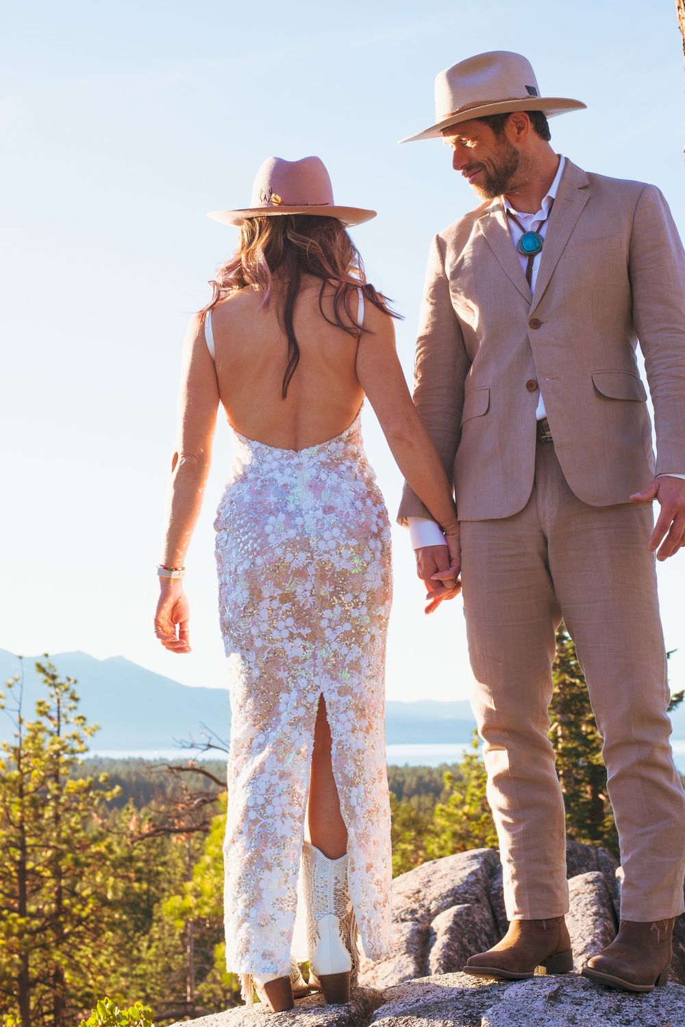 Bride in backless lace gown and groom in tan suit with cowboy hats on rocky overlook.