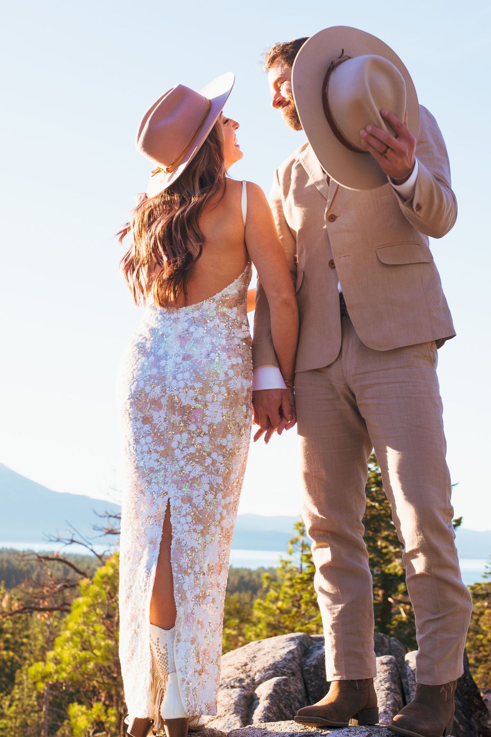 Bride and groom in cowboy hats gazing at each other on rocky cliff at sunset.
